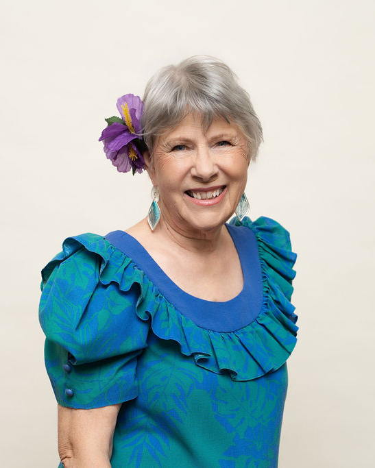 Arlyn J. Macdonald seated at a table wearing a Hawaiian shirt with green leaves. Her head is resting on her right hand. 